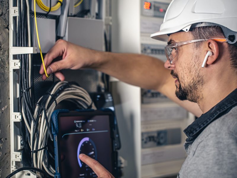 Man, an electrical technician working in a switchboard with fuses. Installation and connection of electrical equipment. Professional uses a tablet.