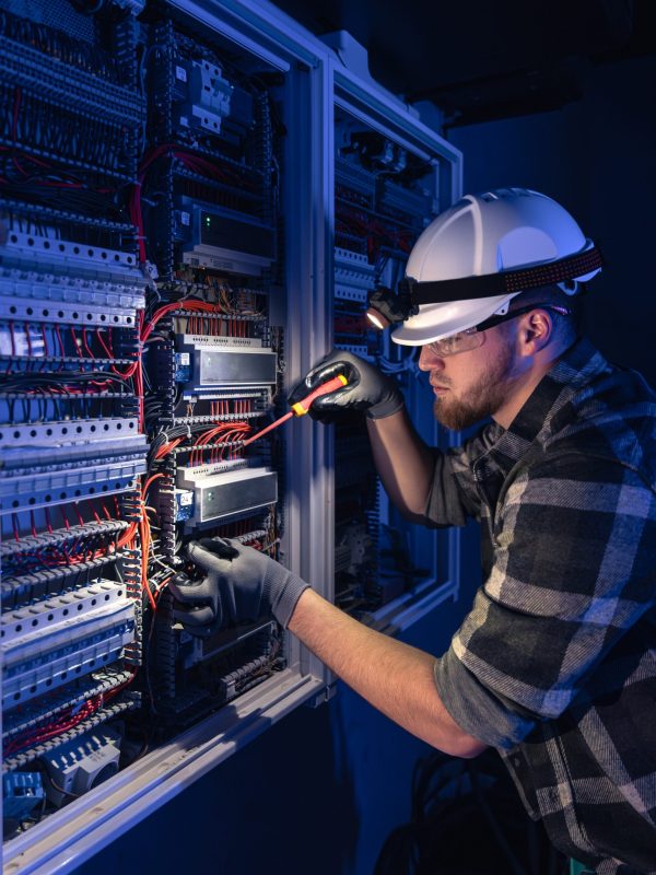 Male electrician working in switchboard with electrical connecting cable. Young adult electrical engineer in special clothes with flashlight on helmet in dark room with emergency lights in background.